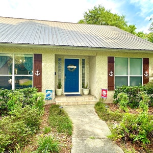 May include: A white house with a blue door and brown shutters. The door has a wreath on it and the shutters have anchors on them. There are plants in pots on either side of the door. The house is surrounded by green bushes.