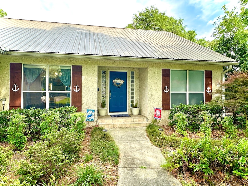 May include: A white house with a blue door and brown shutters. The door has a wreath on it and the shutters have anchors on them. There are plants in pots on either side of the door. The house is surrounded by green bushes.