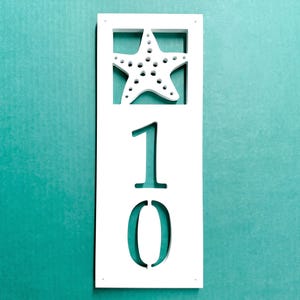 May include: White rectangular house number sign with a starfish cutout at the top and the number 10 cut out in the center. The sign is against a teal background.