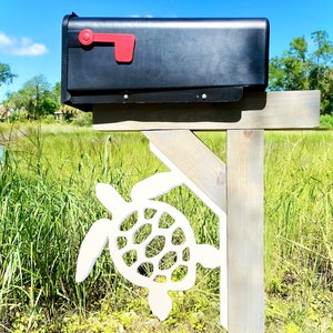 May include: A black mailbox with a red flag is mounted on a wooden post with a white sea turtle cutout. The mailbox is in front of a grassy field.