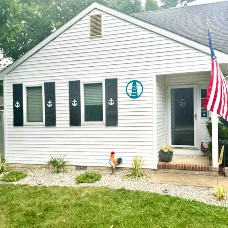 May include: A white house with black shutters and a blue lighthouse sign. The house has a front door with a glass panel and a small porch. There is a red rooster statue in the front yard.