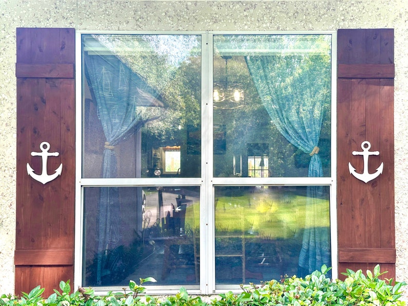 May include: A window with brown wooden shutters and white anchors on each side. The window has a white frame and is divided into four panes. The window is covered with a blue curtain.