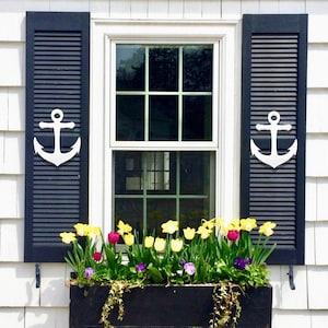 May include: A white window with black shutters and white anchor accents. A window box planter with yellow, pink, and purple flowers is below the window.
