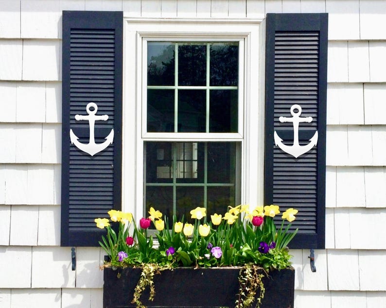 May include: A white window with black shutters and white anchor accents. A window box planter with yellow, pink, and purple flowers is below the window.