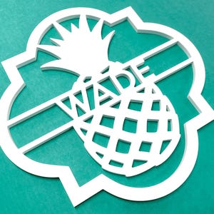 Może przedstawiać: Biały drewniany znak w kształcie ananasa z wyciętym w środku imieniem "Wade". Znak ma ozdobną, falistą ramkę.