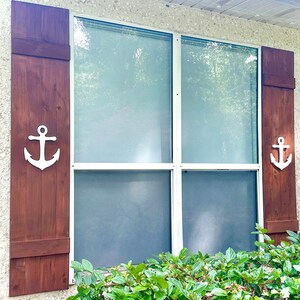 May include: A brown wooden window shutter with a white anchor cutout. The shutter is closed and has a white trim around the edges. The shutter is attached to a white window frame.