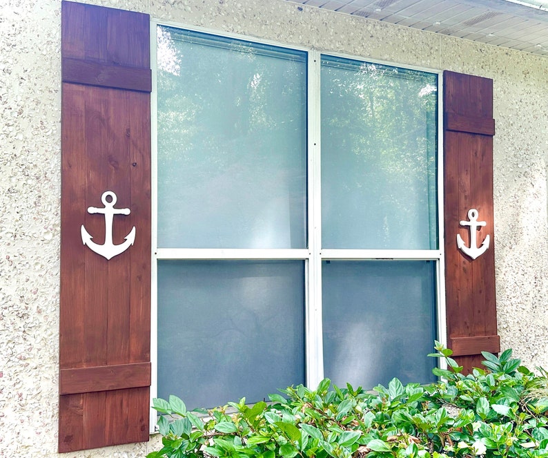 May include: A brown wooden window shutter with a white anchor cutout. The shutter is closed and has a white trim around the edges. The shutter is attached to a white window frame.
