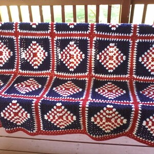 Pu&ograve; includere: Una coperta all'uncinetto in rosso, bianco e blu con un motivo a rombi. La coperta &egrave; fatta di quadrati ed &egrave; drappeggiata su una panca di legno.