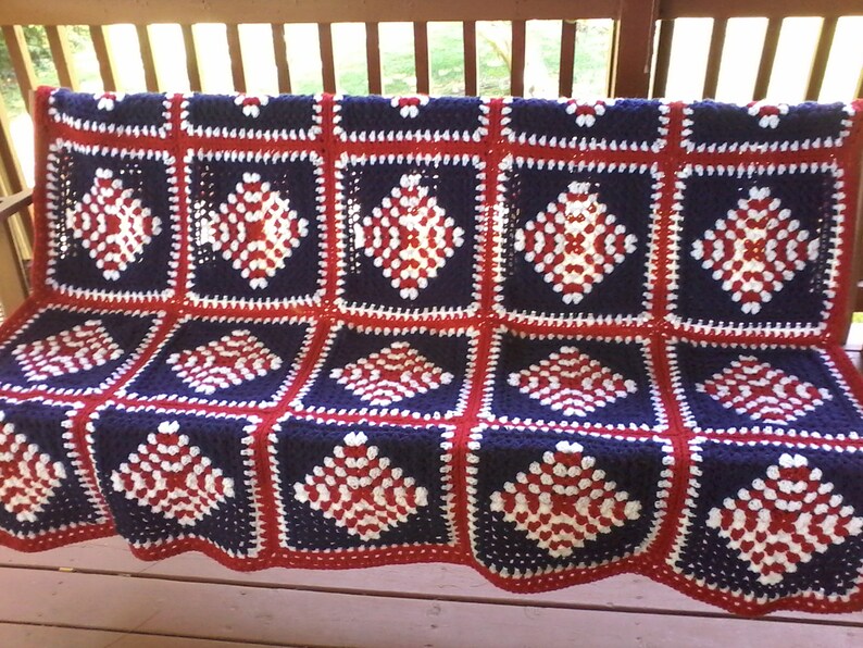 Pu&ograve; includere: Una coperta all'uncinetto in rosso, bianco e blu con un motivo a rombi. La coperta &egrave; fatta di quadrati ed &egrave; drappeggiata su una panca di legno.