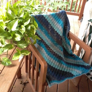 May include: A handmade crochet blanket in shades of blue, teal, and gray rests on a wooden chair. The blanket features a textured stitch pattern and is draped over the chair's seat and back. The chair is on a wooden deck with green plants in the background.