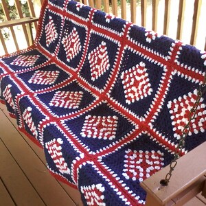 Pu&ograve; includere: Una coperta all'uncinetto in rosso, bianco e blu con un motivo a rombi. La coperta &egrave; drappeggiata su una sedia a dondolo in legno per il portico.