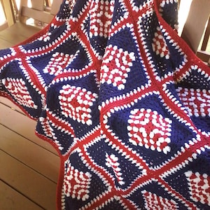 Pu&ograve; includere: Una coperta all'uncinetto in rosso, bianco e blu con un motivo quadrato. La coperta &egrave; realizzata in filato e ha una superficie testurizzata.