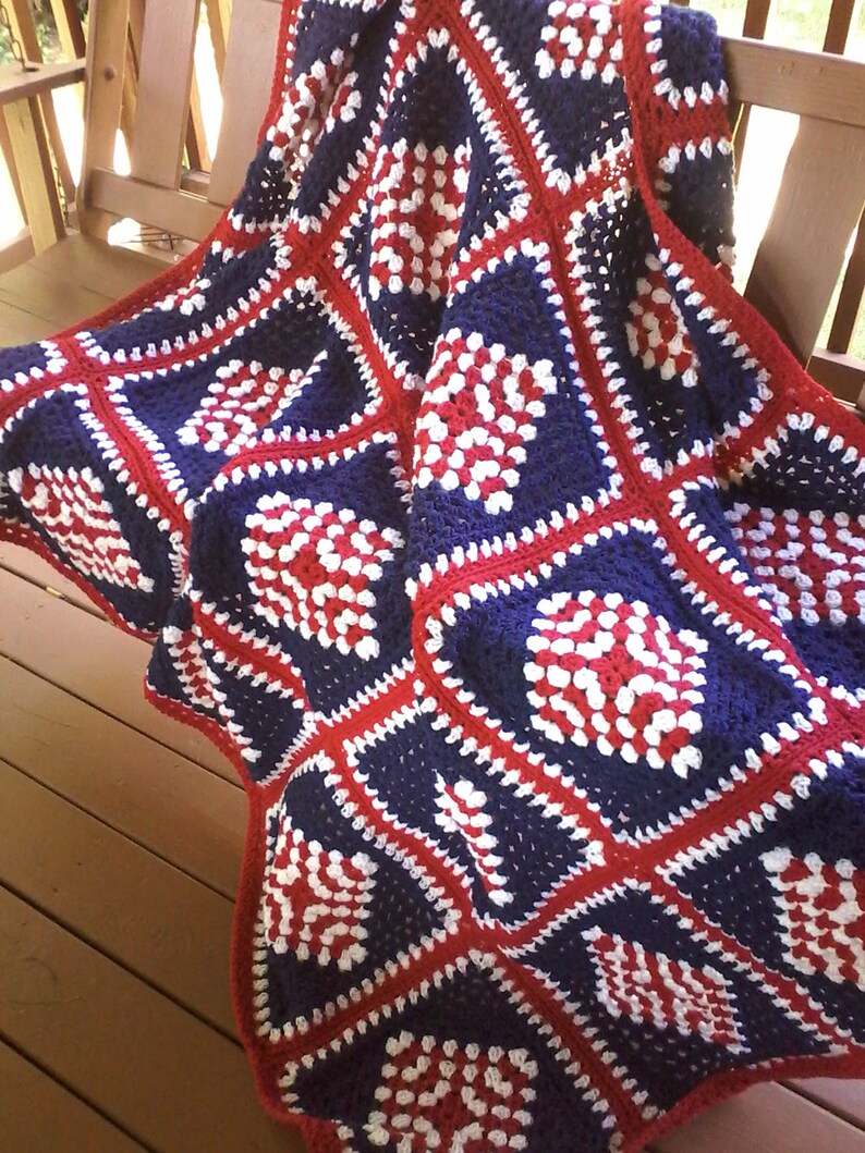 Pu&ograve; includere: Una coperta all'uncinetto in rosso, bianco e blu con un motivo quadrato. La coperta &egrave; realizzata in filato e ha una superficie testurizzata.