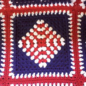 Pu&ograve; includere: Una coperta all'uncinetto con uno schema di colori rosso, bianco e blu. La coperta &egrave; composta da quadrati con un motivo a rombo al centro.