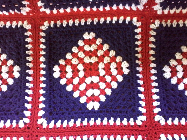 Pu&ograve; includere: Una coperta all'uncinetto con uno schema di colori rosso, bianco e blu. La coperta &egrave; composta da quadrati con un motivo a rombo al centro.
