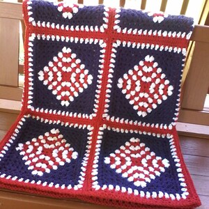 Pu&ograve; includere: Una coperta all'uncinetto con un motivo rosso, bianco e blu. La coperta &egrave; fatta di quadrati con una forma di diamante al centro. La coperta &egrave; piegata e appoggiata su una superficie di legno.