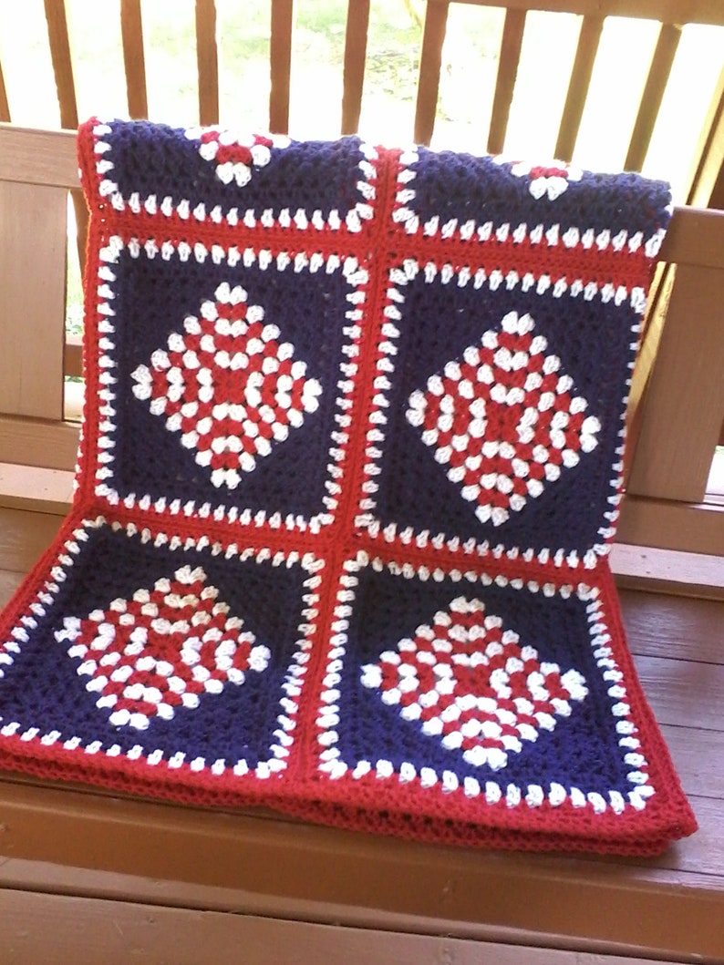 Pu&ograve; includere: Una coperta all'uncinetto con un motivo rosso, bianco e blu. La coperta &egrave; fatta di quadrati con una forma di diamante al centro. La coperta &egrave; piegata e appoggiata su una superficie di legno.