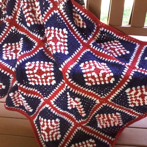 Pu&ograve; includere: Una coperta all'uncinetto in rosso, bianco e blu con un motivo quadrato. La coperta &egrave; piegata e drappeggiata su una ringhiera di veranda in legno.