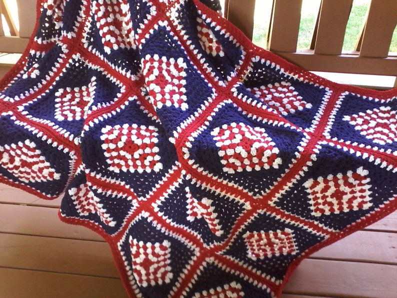 Pu&ograve; includere: Una coperta all'uncinetto in rosso, bianco e blu con un motivo quadrato. La coperta &egrave; piegata e drappeggiata su una ringhiera di veranda in legno.