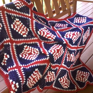 Pu&ograve; includere: Una coperta all'uncinetto in rosso, bianco e blu con un motivo a rombi. La coperta &egrave; realizzata in filato e ha un bordo a frange.