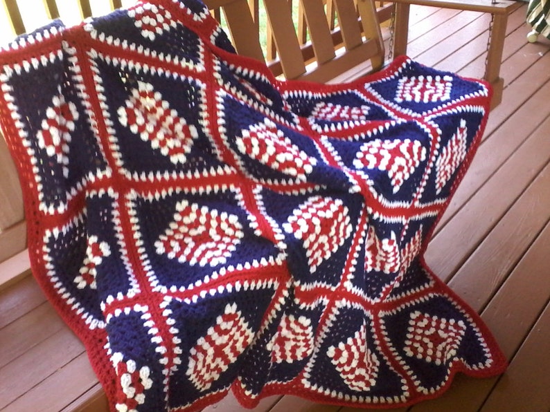 Pu&ograve; includere: Una coperta all'uncinetto in rosso, bianco e blu con un motivo a rombi. La coperta &egrave; realizzata in filato e ha un bordo a frange.