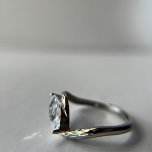 May include: A silver ring with a pear-shaped, clear gemstone. The band has a textured design on one side and a smooth, curved design on the other, creating a modern, elegant look. The gemstone is set in a prong setting.