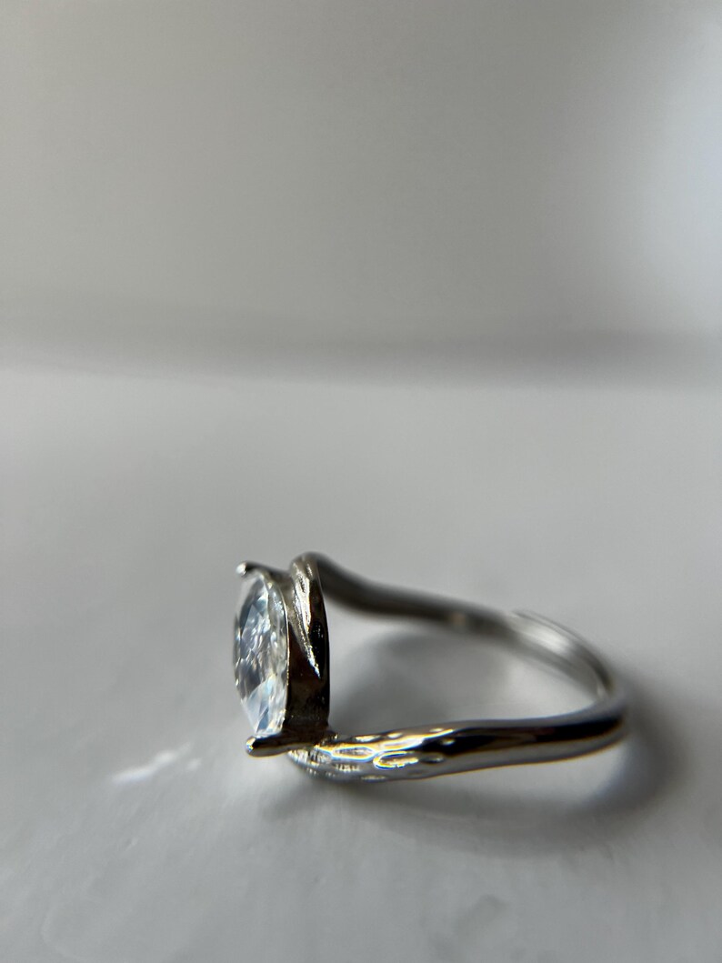 May include: A silver ring with a pear-shaped, clear gemstone. The band has a textured design on one side and a smooth, curved design on the other, creating a modern, elegant look. The gemstone is set in a prong setting.
