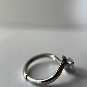 May include: A silver ring with a single, square-cut, clear gemstone. The ring band is smooth and rounded, with a slight textured detail near the gemstone setting. The ring is set against a white background.