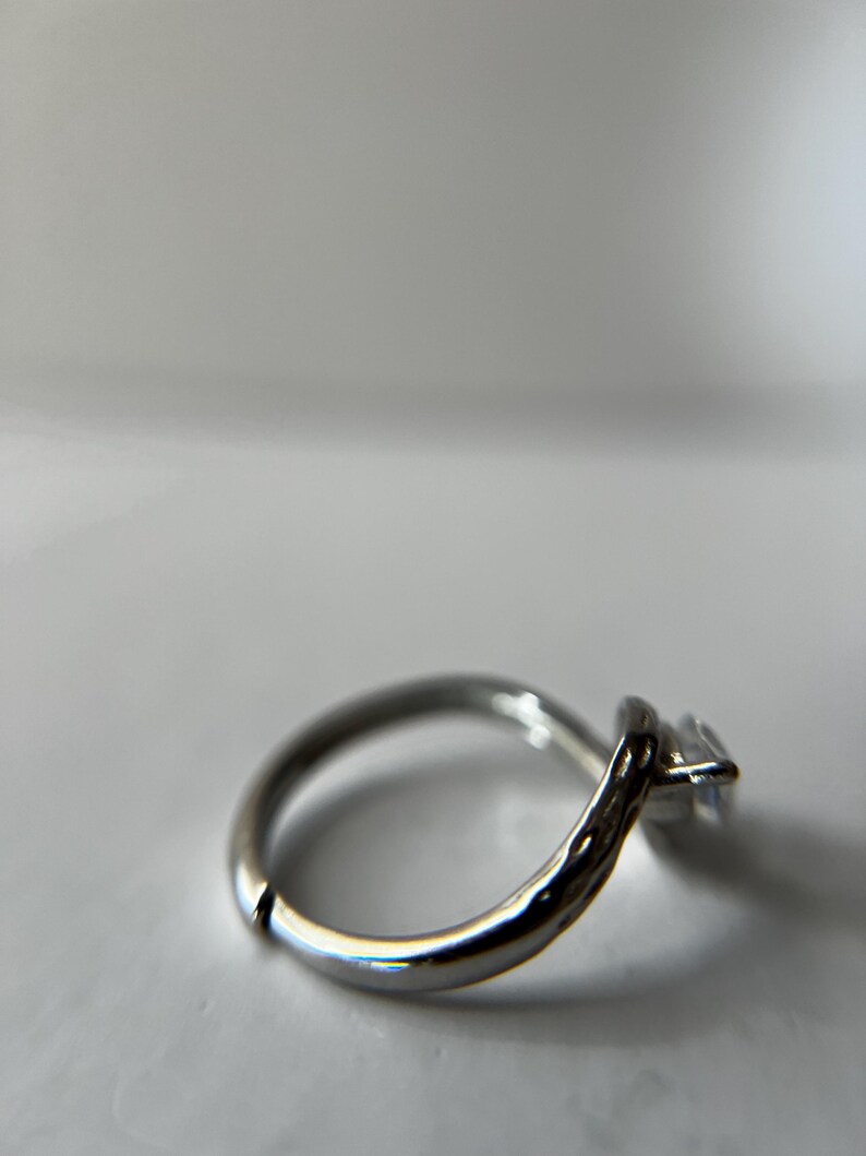 May include: A silver ring with a single, square-cut, clear gemstone. The ring band is smooth and rounded, with a slight textured detail near the gemstone setting. The ring is set against a white background.