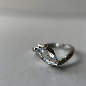 May include: A silver ring with a marquise-cut, clear gemstone. The band has a textured, twisted design. The gemstone is set in a prong setting, and the ring is displayed on a light gray surface.