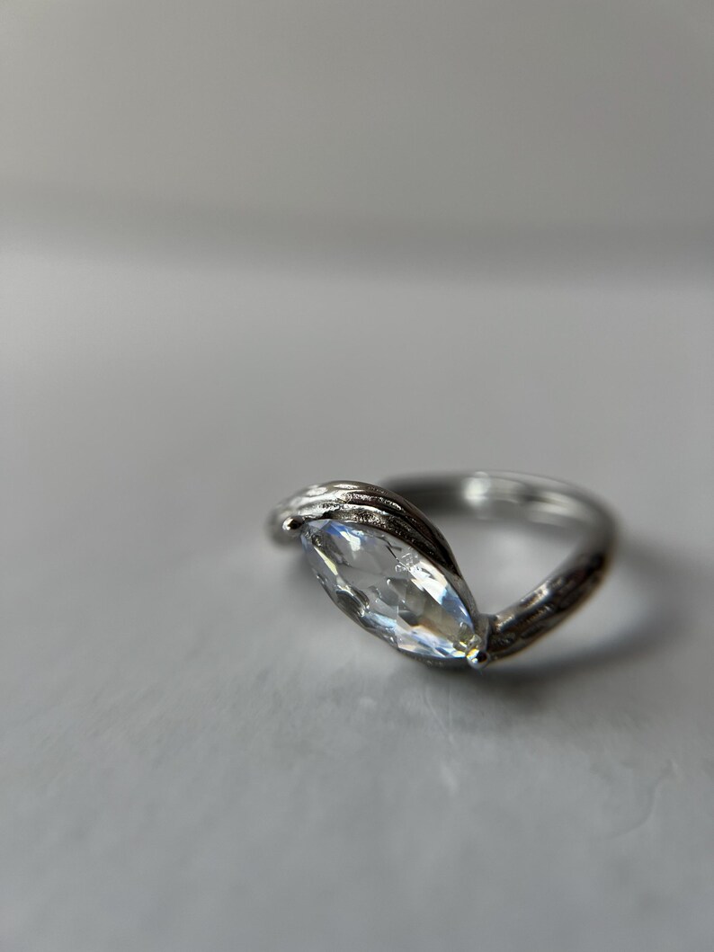 May include: A silver ring with a marquise-cut, clear gemstone. The band has a textured, twisted design. The gemstone is set in a prong setting, and the ring is displayed on a light gray surface.