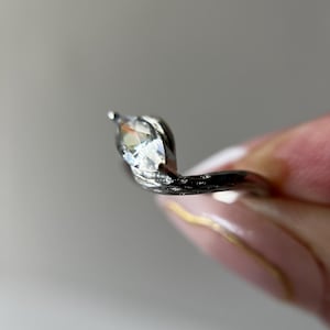 May include: A silver ring with a marquise-cut gemstone. The ring's band has a textured, organic design. The gemstone is clear and faceted, catching the light. The ring is shown against a soft, neutral background.