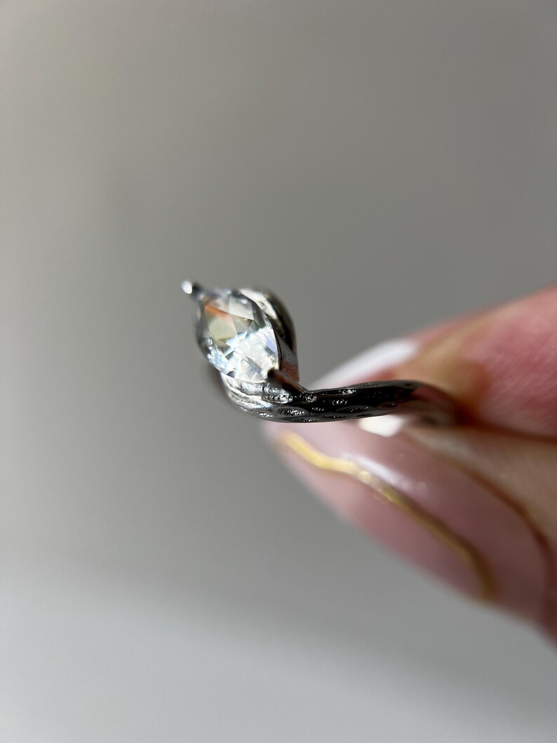 May include: A silver ring with a marquise-cut gemstone. The ring's band has a textured, organic design. The gemstone is clear and faceted, catching the light. The ring is shown against a soft, neutral background.