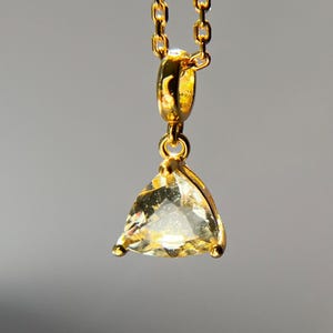 May include: A gold necklace featuring a triangular, pale yellow gemstone pendant. The pendant is set in a gold frame and suspended from a fine gold chain. The backdrop is a muted grey.