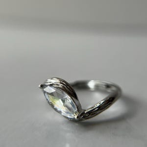 May include: A silver ring with a marquise-cut clear gemstone. The band has a textured, vine-like design. The gemstone is set in a V-shaped setting, creating a unique and elegant look.