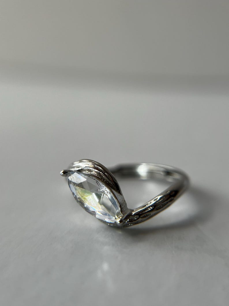 May include: A silver ring with a marquise-cut clear gemstone. The band has a textured, vine-like design. The gemstone is set in a V-shaped setting, creating a unique and elegant look.