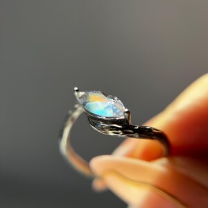 May include: A silver ring with a marquise-cut gemstone. The gemstone has a rainbow iridescence. The ring's band has a textured, branch-like design. The ring is held against a blurred background.
