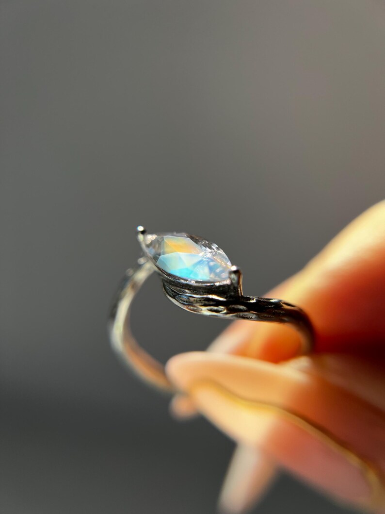 May include: A silver ring with a marquise-cut gemstone. The gemstone has a rainbow iridescence. The ring's band has a textured, branch-like design. The ring is held against a blurred background.