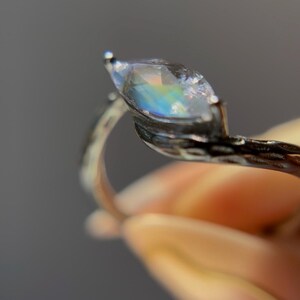 May include: A silver ring featuring a marquise-cut gemstone with iridescent rainbow hues. The ring's band has a textured, organic design. The gemstone is held in place by delicate prongs, creating a unique and elegant piece of jewelry.
