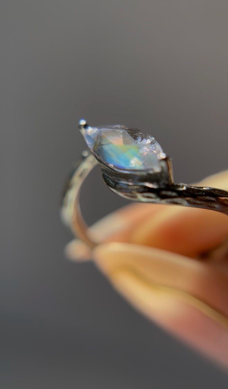 May include: A silver ring featuring a marquise-cut gemstone with iridescent rainbow hues. The ring's band has a textured, organic design. The gemstone is held in place by delicate prongs, creating a unique and elegant piece of jewelry.