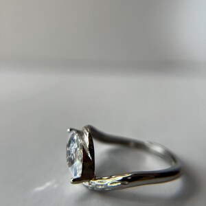 May include: A silver ring with a pear-shaped, clear gemstone. The band curves around the finger, with the gemstone held in place by two prongs. The ring is set against a white background.