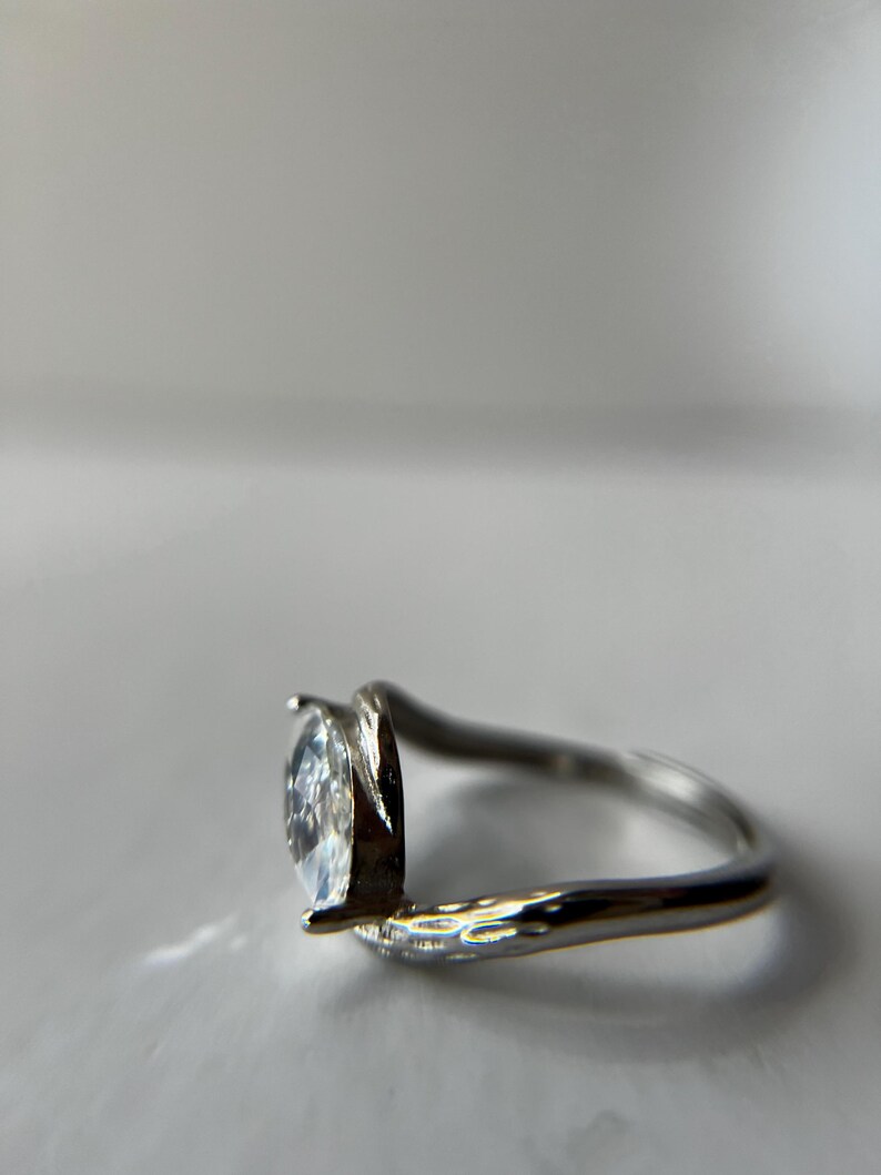 May include: A silver ring with a pear-shaped, clear gemstone. The band curves around the finger, with the gemstone held in place by two prongs. The ring is set against a white background.