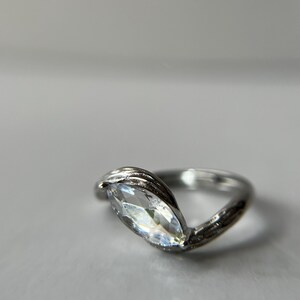 May include: A silver ring with a marquise-cut, clear gemstone. The band has a textured, leaf-like design that wraps around the gemstone. The ring is set against a soft, neutral background.