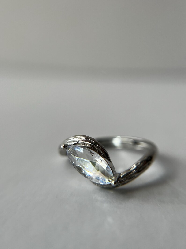 May include: A silver ring with a marquise-cut, clear gemstone. The band has a textured, leaf-like design that wraps around the gemstone. The ring is set against a soft, neutral background.