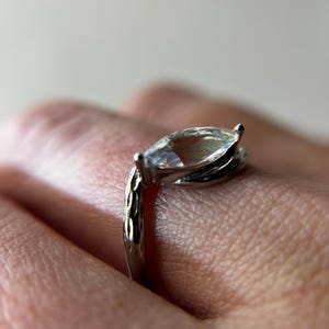 May include: A silver ring with a marquise-cut gemstone. The ring's band has a textured design, and the gemstone is held in place by prongs. The ring is shown on a person's finger.