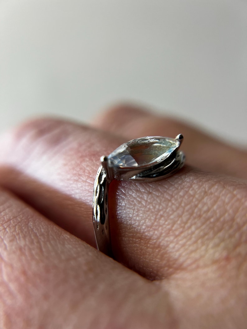 May include: A silver ring with a marquise-cut gemstone. The ring's band has a textured design, and the gemstone is held in place by prongs. The ring is shown on a person's finger.