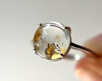 May include: A clear, round gemstone ring with a silver band. The gemstone features inclusions of yellow and brown, resembling a miniature landscape. The ring is held up against a white background, showcasing the details of the jewelry.