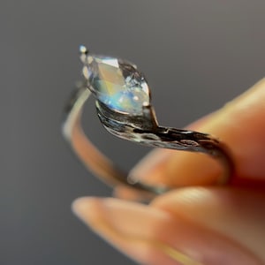 May include: A marquise-cut gemstone ring with a silver band. The gemstone reflects a rainbow of colors. The band has a textured, organic design. The ring is held up against a gray background.