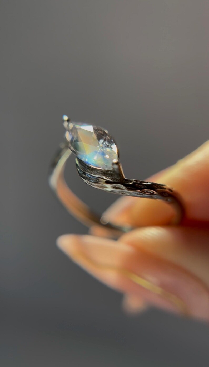 May include: A marquise-cut gemstone ring with a silver band. The gemstone reflects a rainbow of colors. The band has a textured, organic design. The ring is held up against a gray background.