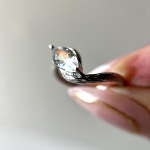 May include: A silver ring with a marquise-cut gemstone. The ring has a unique, swirling design with small accent stones. The gemstone is clear and reflects light, creating a sparkling effect. The ring is held by a person.